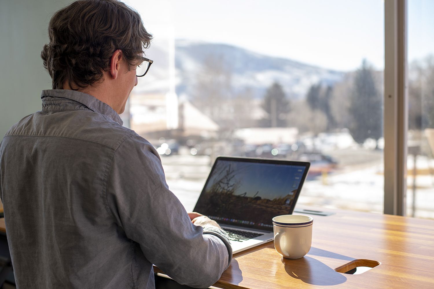 A man works at his computer at a sunny window with mountains outside.