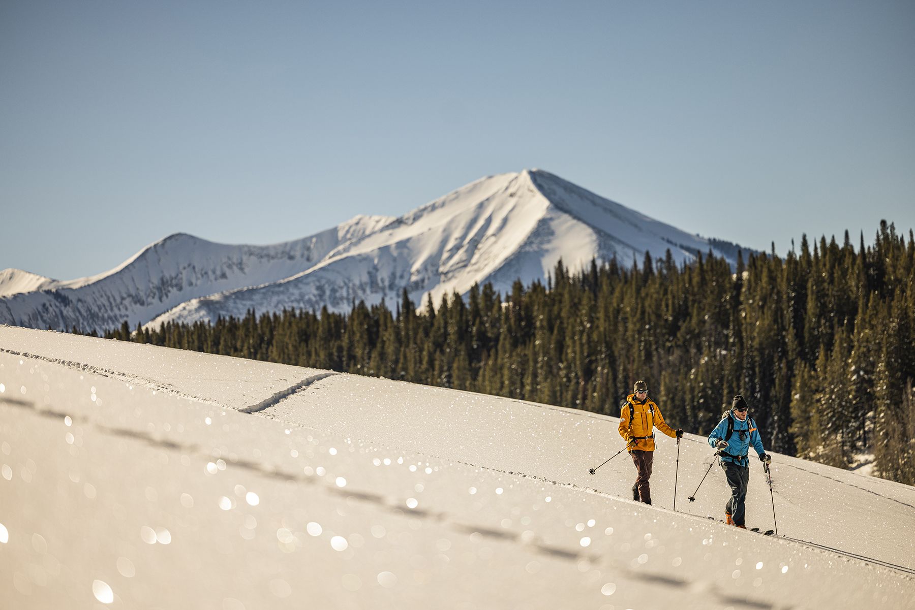 Two backcountry skiers skin up a gentle slope where the snow is covered in sparkly hoar frost. A snow-covered peak towers in the background.
