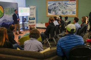 A group of people sitting in rows of chairs watching a presentation in a room with a picture of a map, teal wall, and materials for a presentation