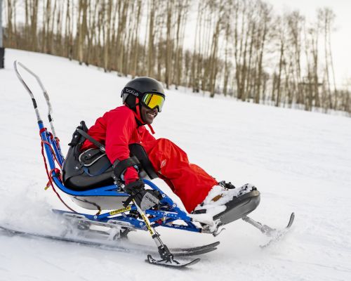 A boy on an adaptive ski with the adaptive sports center in Crested Butte. The ski has a seat on it with two handles that have runners. There are runners under the seat as well. The boy's feet are propped up on a platform. The boy is also wearing a helmet and googles.