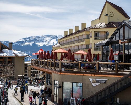 butte 66 crested butte mountain resort. a restaurant with a large deck and tables with umbrellas overlooks a concrete patio and the base of a ski area.