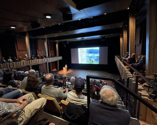 crested butte public policy forum talk. people sit in a theater while someone speaks from a podium on a stage.