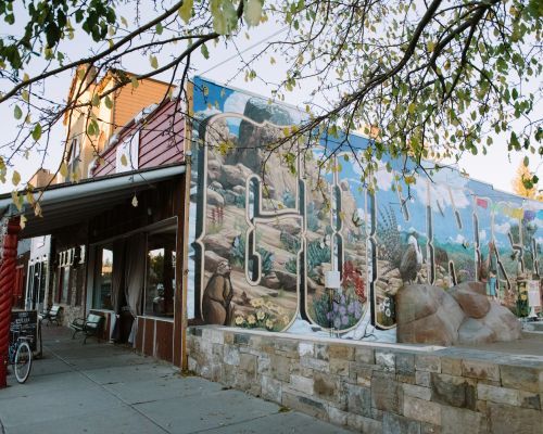 the outside of a building with a large, colorful mural that says "Gunnison" and depicts scenes of outdoor recreation and flora and fauna. The front of the building has a red awning and a sign that says "1880 tapas and spirits." Big windows and a door are under the awning