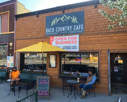 The outside of Back Country Cafe. A sign above the windows and doors reads "Back County Cafe Breakfast - Lunch - Cocktails." Two cafe tables sit under an umbrella where two men are sitting and talking.