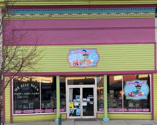 A storefront that features big windows with games and books. One window has the words "The Book Worm Books * Games * Maps." The other window says "Abracadabra" and features a caterpillar wearing glasses and sitting on top of a stack of books surrounded by blocks, a teddy bear, and a train. The bottoms of the windows say "Gifts, Candles, Cards, Books, Journals, Games, Toys, Puzzles, Colorado Gifts." The caterpillar logo is above the door into the store.