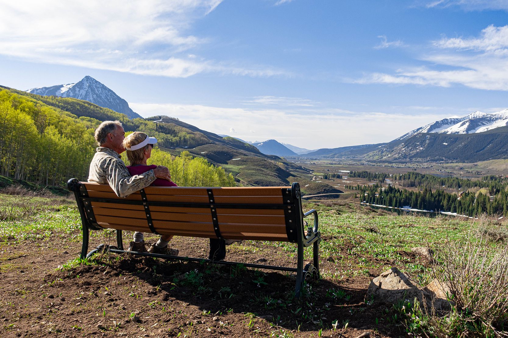 A couple sits on a trailside bench on a mostly sunny spring day in Crested Butte.