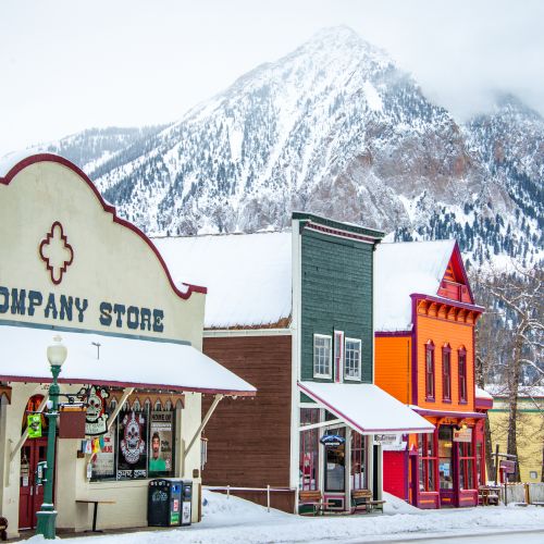 A downtown street covered in snow with a snowcapped mountain peak in the background. This is Elk Avenue in winter in Crested Butte