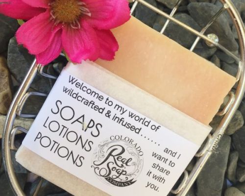 A bar of soap in a soap dish next to a flower. The label says "Welcome to my world of wildcrafted & infused......Soaps, Lotions, Potions. And I want to share it with you." A circular logo that says "Colorado Real Soap Company" also rests on the logo.