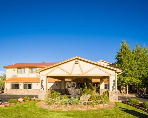 Alpine Inn in Gunnison. The hotel is two stories. There is an overhang roof area above the pull through to unload baggage and check-in. The overhang has a silhouette of a cowboy and a cowgirl, one on each column. A statue of a bighorn sheep stands in a garden in front of the hotel.