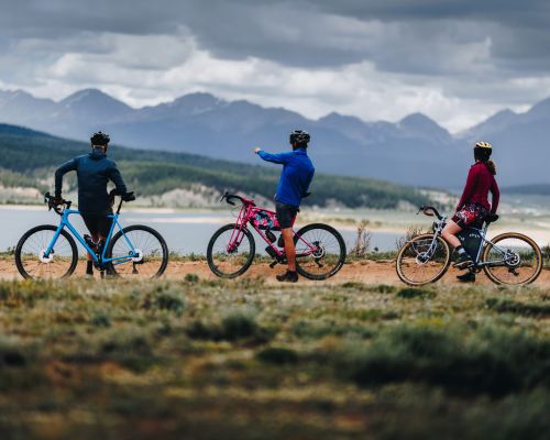 Three people on bikes face a lake with mountain peaks in the background