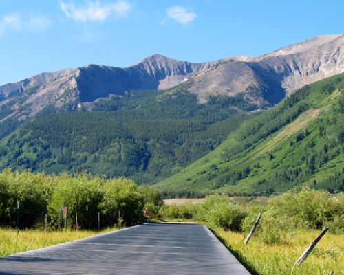 A paved path cutting through a grassy meadow with mountain peaks in the background.