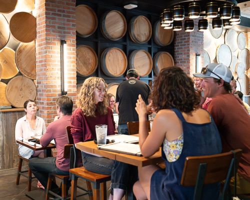 the interior of Bruhaus Crested Butte. Men and women sit at high and low top tables while talking. Barrels line the walls. A light feature with round fixtures hangs from the ceiling.