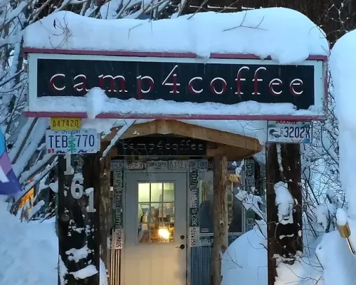 Camp 4 Coffee in Crested Butte, CO. The doorway is topped with snow. A sign that reads "Camp 4 Coffee" sits above the door. Warm, cozy light shines from inside the door.