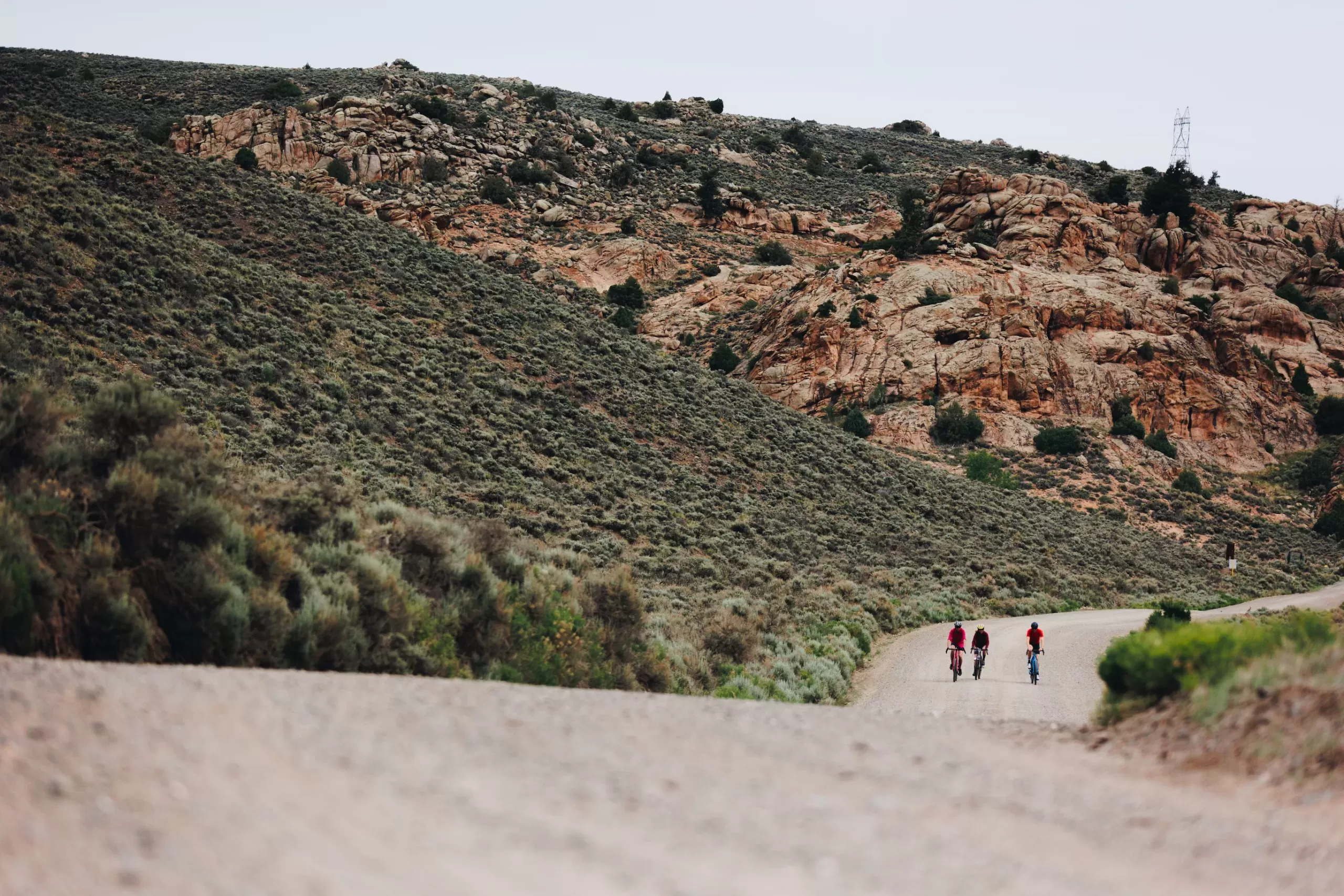 Three people on bikes ride on a gravel road with rocks in the background