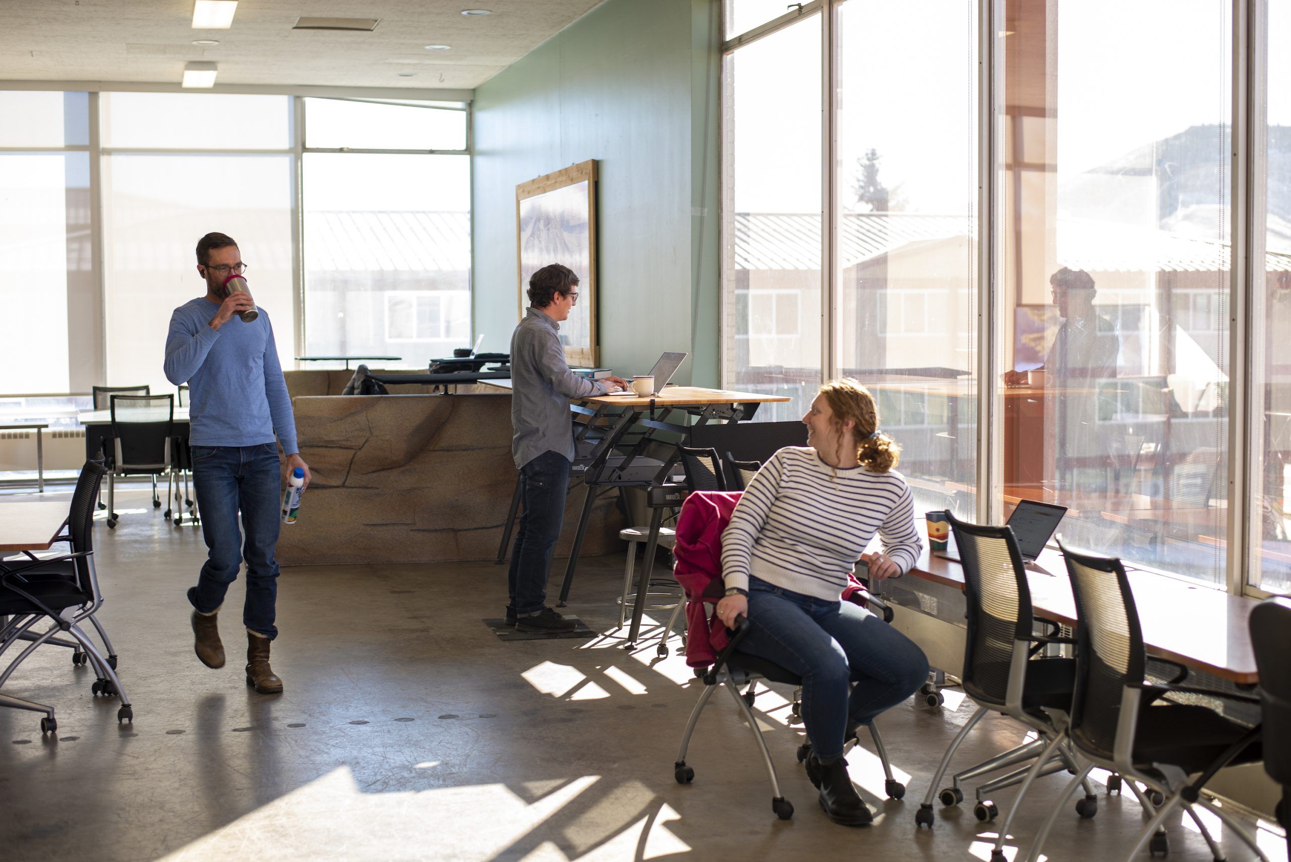 One person walks, one person stands at a desk, and one person sits at a desk in a workspace