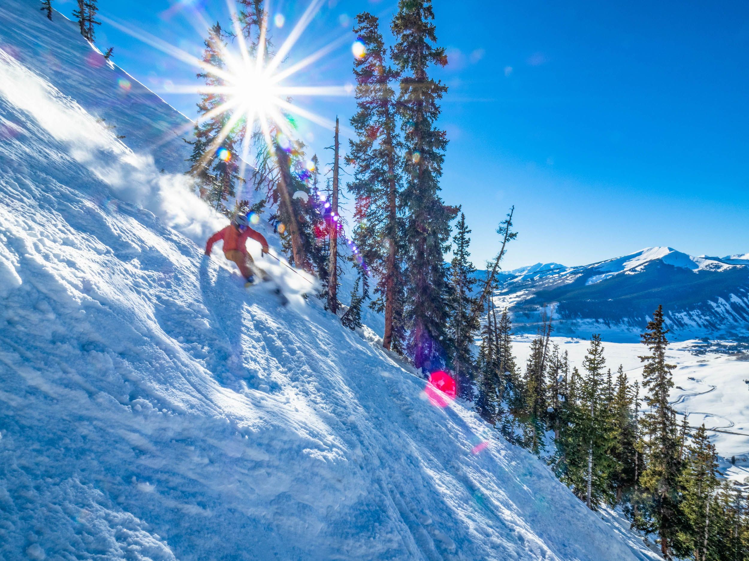 A skier rides down a steep slope with trees and mountain peaks in the background.