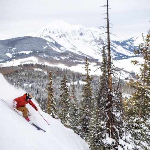 A person skis down a steep slope with a mountain peak in the background. Crested Butte Mountain Resort is known for steep skiing.