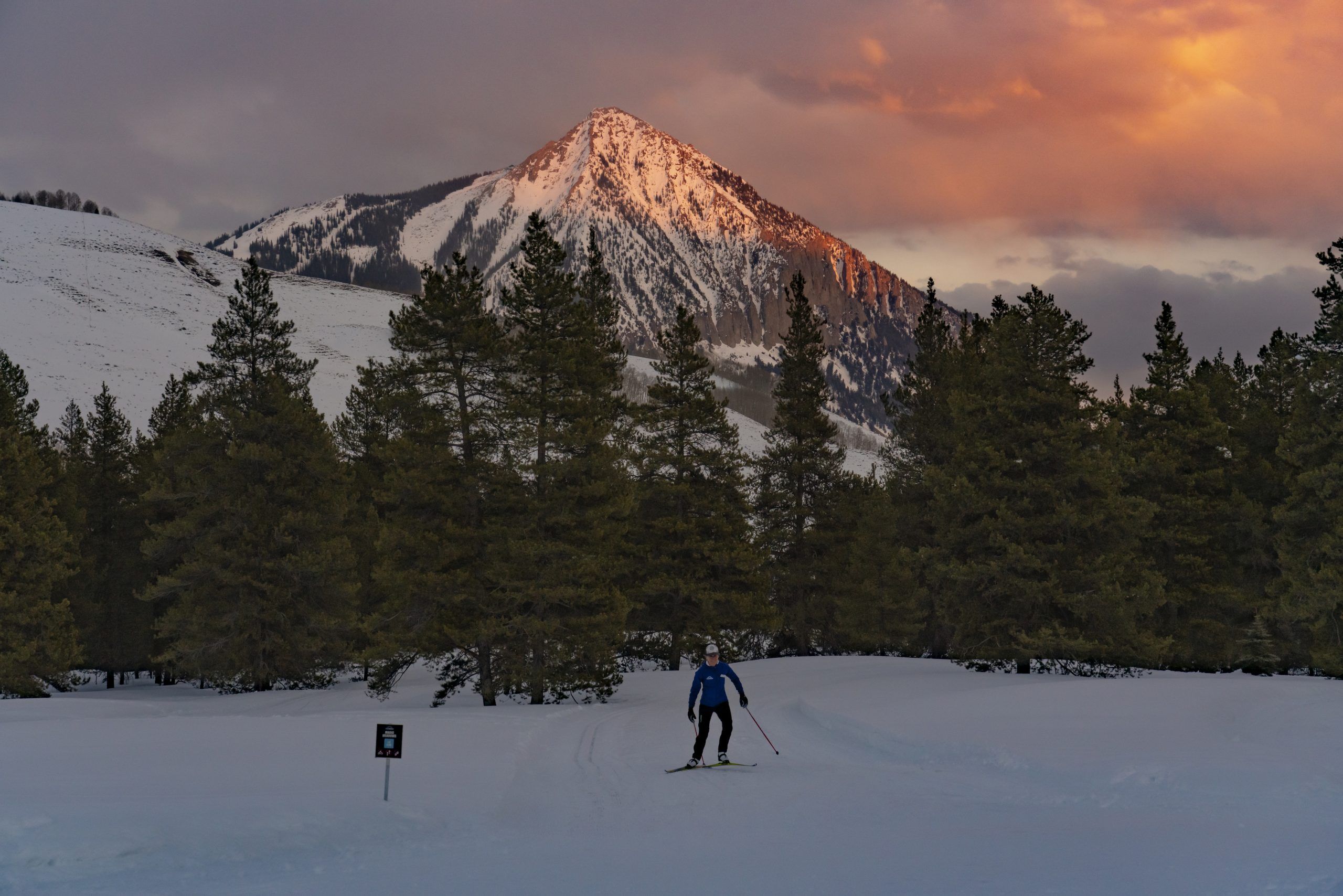 A person cross-country skis on a snowy path under a sunset and a mountain peak