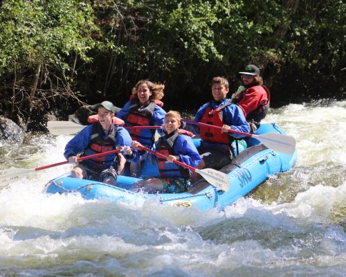 A guide and a family of four paddle through a wave in the river.