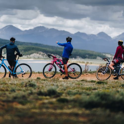 Three people on bikes face a lake with mountain peaks in the background