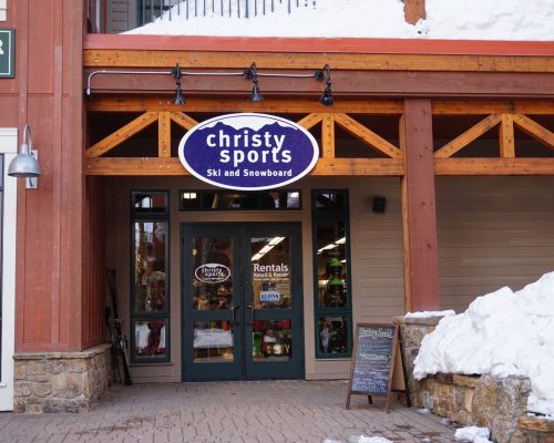The storefront for Christy Sports. The double doors feature a logo that says "Christy Sports" and the hours for the store. A round sign above the door reads "Christy Sports Ski and Snowboard" with an outline of a mountain peak.