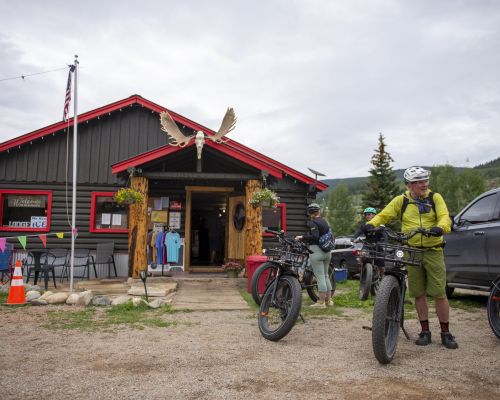 People ride bikes in front of a lodge-style building with a pair of moose antlers over the door. This is the stumbling moose lodge in pitkin, colorado