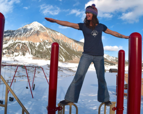 A woman with the crested butte yoga co-op balancing on playground equipment in Crested Butte. Crested Butte Mountain, a peak that comes to a point, is in the background of the image.