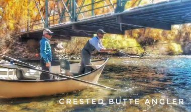 The exterior for Crested Butte Anglers. A sign rests above the windows. It reads "Crested Butte Angler Guide Service * Fly Shop * Outdoor Apparel * Gifts" with a jumping fish. The windows have decals that say "outdoor apparel," "guide service," "fly shop," gifts" and "t-shirts." A log bench sits in front of the store.