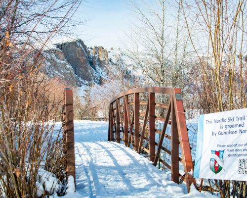 A sign that says "This Nordic ski trail system is groomed by Gunnison Nordic" in front of a bridge with rock formations in the background