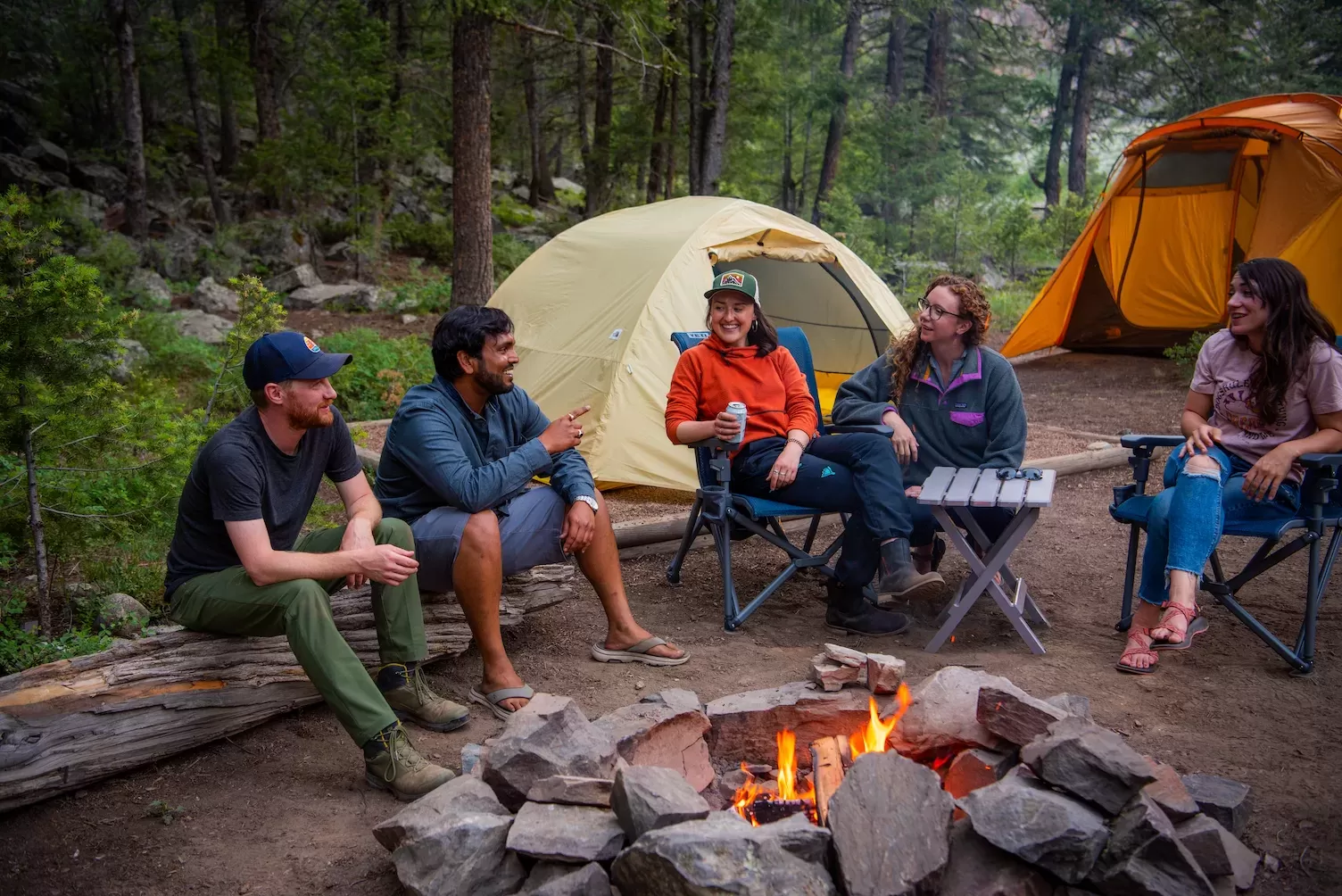 Five people sit around a campfire. There are tents set up in the background