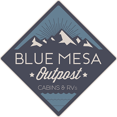 Two cabins at Blue Mesa Outpost. A car and picnic table are next to the cabins. A lake and mountain peaks are in front of the cabins.