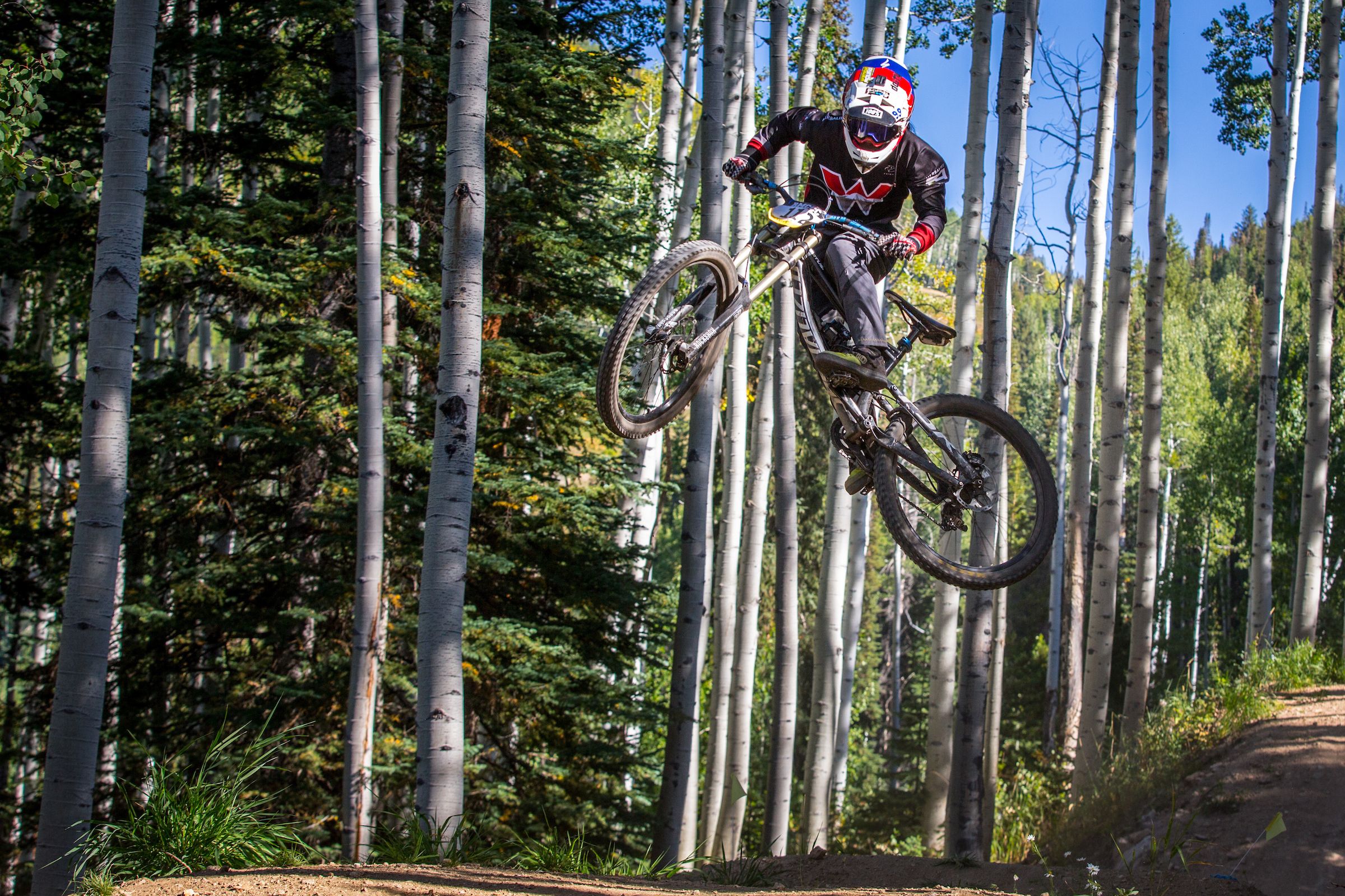 A mountain biker in a Western Colorado University jersey jumps on a mountain bike on a trail through aspen trees.