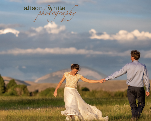 A man and a woman pose for a photo with alison white photography. Fields, trees and a rounded mountain top under a cloudy sky are in the background. A watermark that says "Alison White Photography" sits at the top of the lefthand side of the image