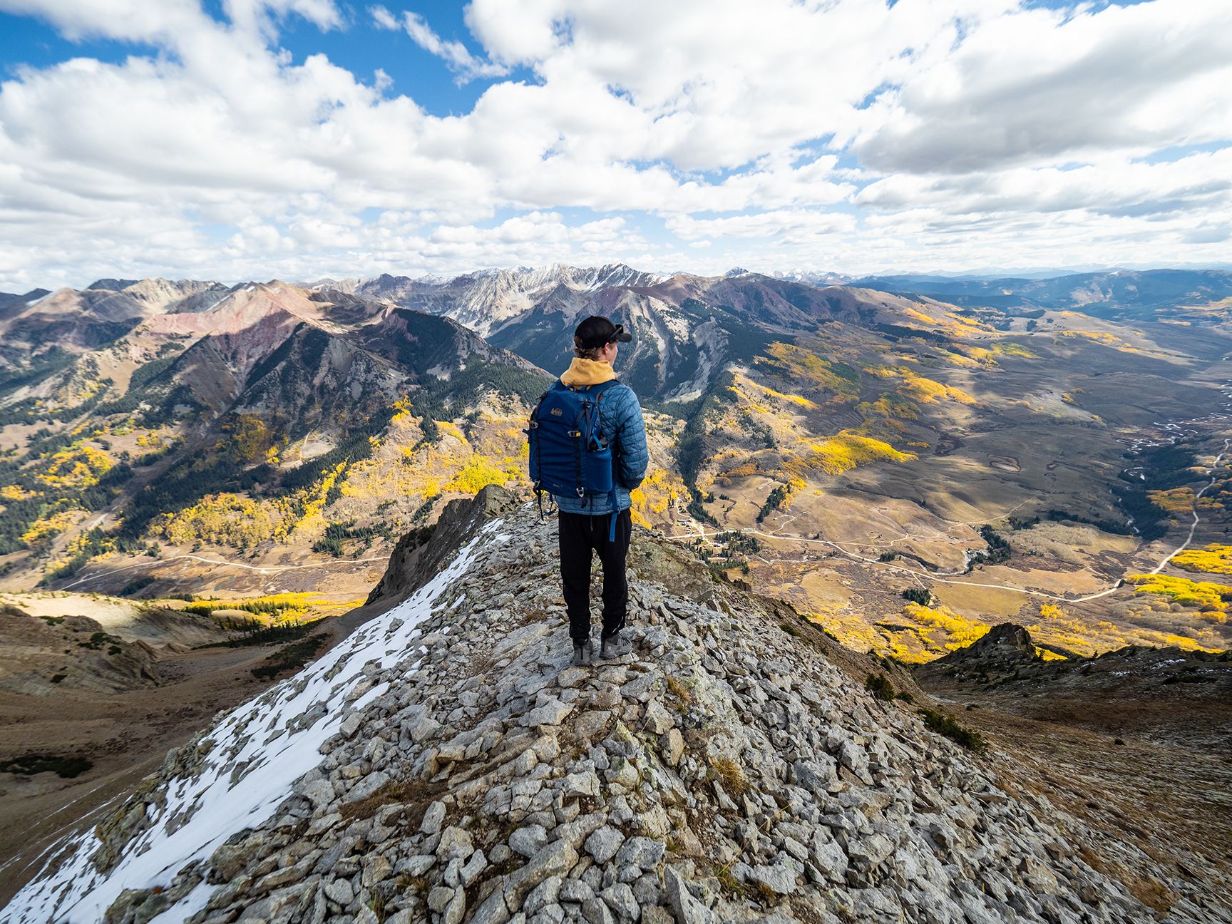 A man hikes on a rocky ridge overlooking a mountain valley with yellow aspen leaves.