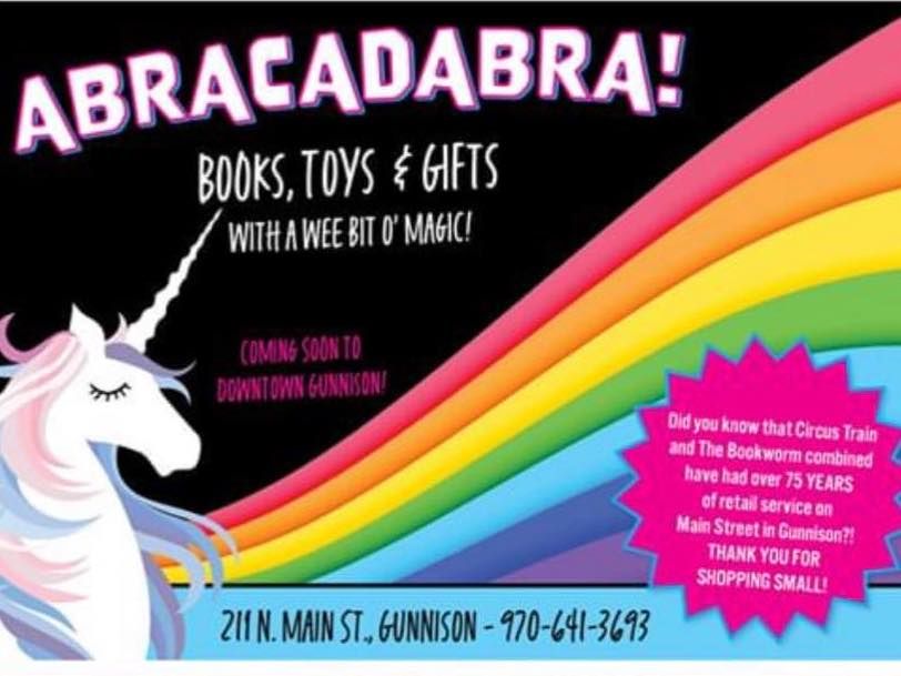 A storefront that features big windows with games and books. One window has the words "The Book Worm Books * Games * Maps." The other window says "Abracadabra" and features a caterpillar wearing glasses and sitting on top of a stack of books surrounded by blocks, a teddy bear, and a train. The bottoms of the windows say "Gifts, Candles, Cards, Books, Journals, Games, Toys, Puzzles, Colorado Gifts." The caterpillar logo is above the door into the store.