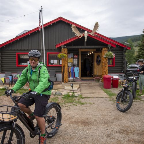 People on bikes in front of a log cabin building with moose antlers mounted above the door. This lodging in pitkin is the stumbling moose lodge