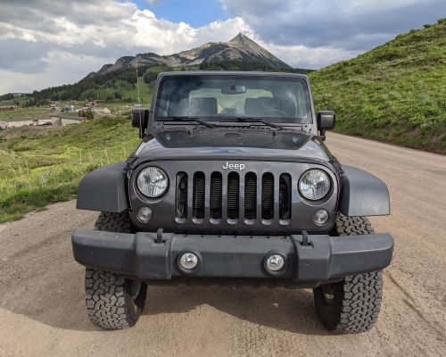 Alpenglow, a jeep rental company in Crested Butte. The picture depicts a Jeep facing the camera head-on. The Jeep is parked on a dirt road with a pointy mountain peak in the background.