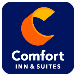 The exterior of the Comfort Inn in Gunnison. The three story building has a porte co·chère above the entrance. Trees and grassy areas are in the parking lot with some parked trucks. There is a large sign in the parking lot that says "Comfort Inn" with a round, striped logo.