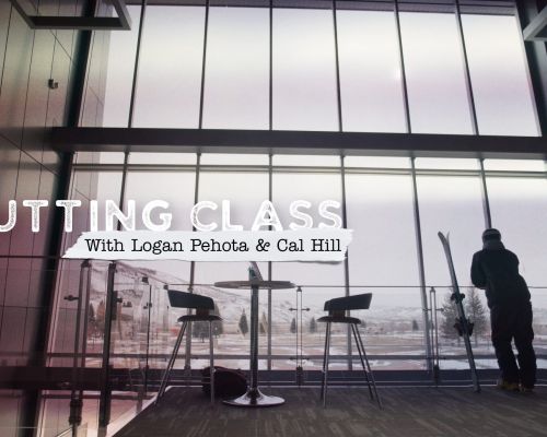 A student leans up against the window of a university building, looking outside. He is dressed in ski gear, with his skis leaning beside him.