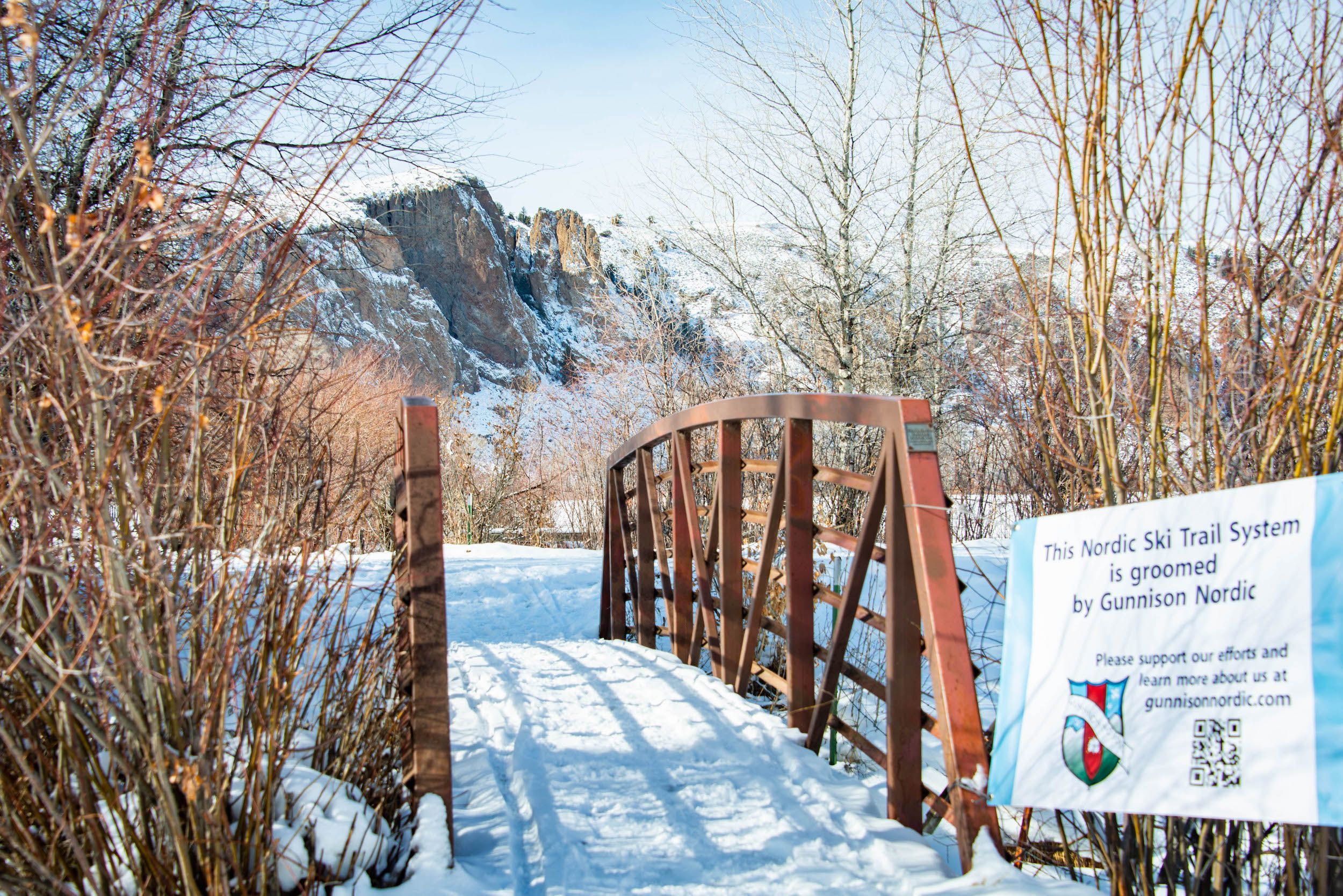 a bridge with a sign in front of it that says "This Nordic Ski Trail System is groomed by Gunnison Nordic." Land stewardship