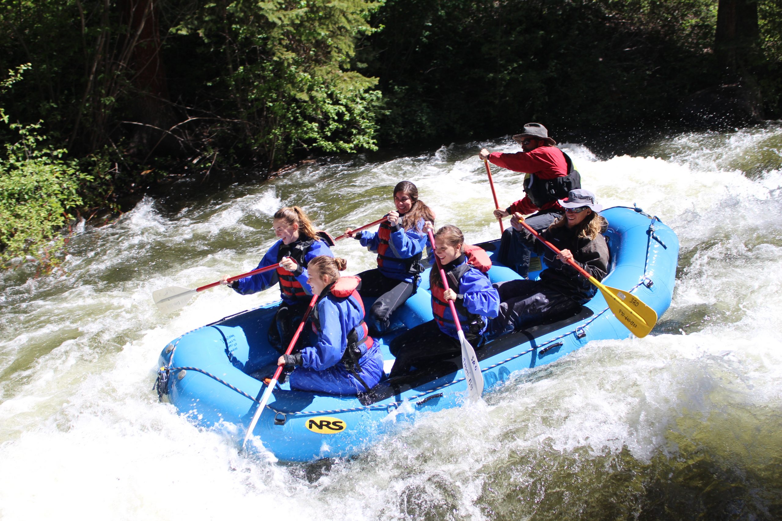 Crested Butte Commercial Whitewater Rafting Six people paddle a blue raft through a large wave on a river.