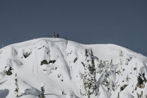 A stock photo for the Natural Selection tour. Snowboarders stand on a snowy ridge overlooking steep terrain.