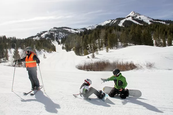 An adaptive snowboarder, employee and a volunteer take a break on a ski run in front of a mountain peak