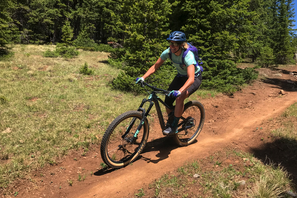 A smiling female mountain biker rides down a flat dirt trail through spruce trees in Crested Butte