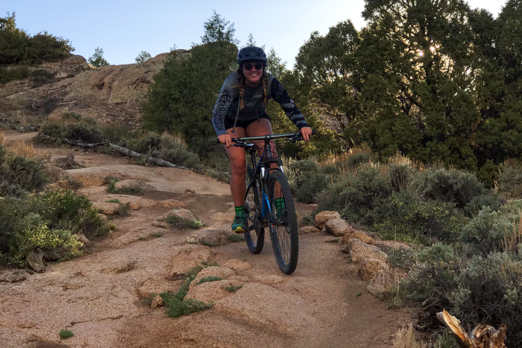 Crested Butte mountain biking female biker smiles as she rides downhill on a sandstone trail. The area is a high desert in summer.