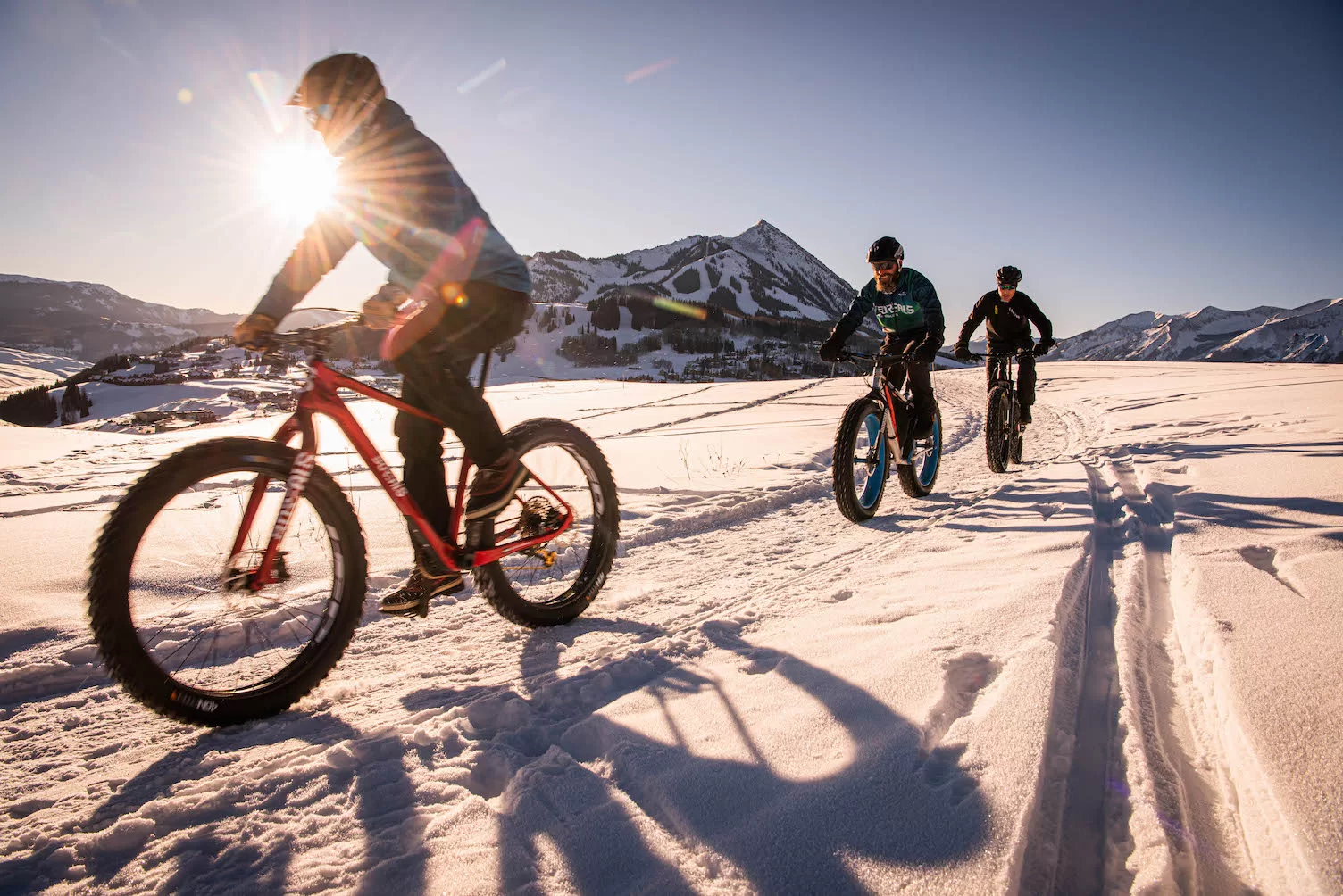 Three fat bikers ride a groomed track. The bikes have wide tires. There is a mountain peak in the background