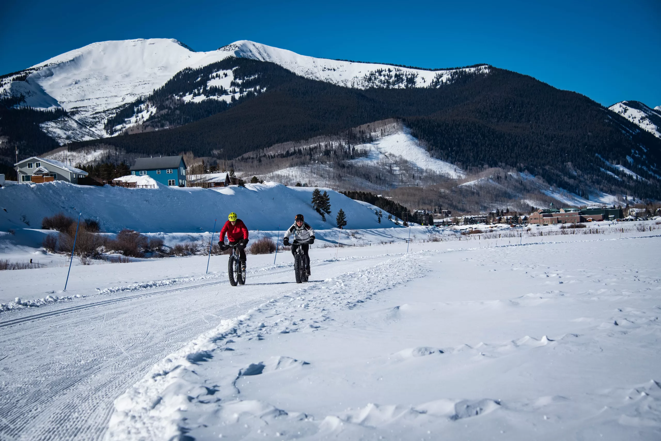 Two people ride fat bikes, which are special bikes designed for snow, on a groomed track. A mountain peak is in the background.