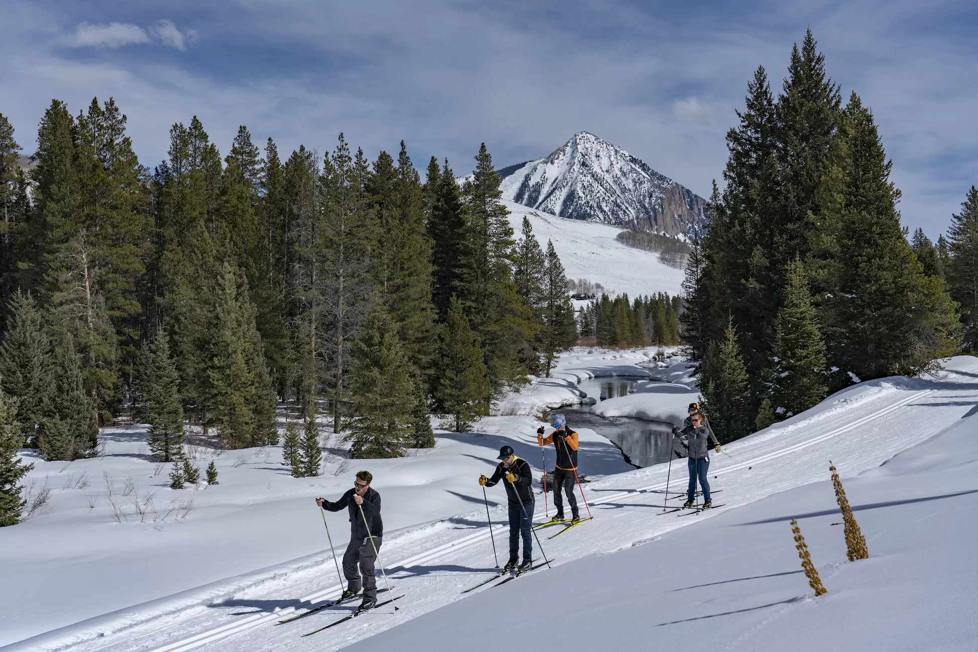 Four people on nordic skis ski on a groomed snow track. There is a mountain peak in the background.