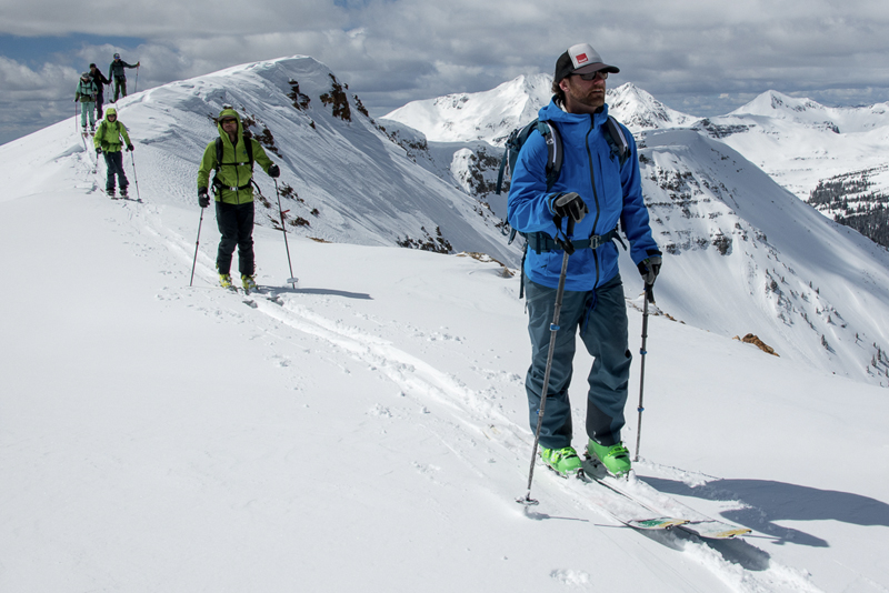 Six skiers skin up a snowy ridge with steep, rocky mountains in the background.