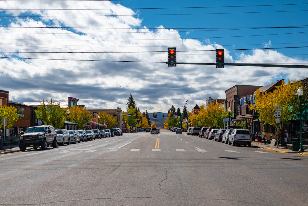 A wide shot of a downtown street with businesses on each side. The street is lined with trees where the leaves are turning colors and cars. There is a stoplight and the sky is blue and cloudy